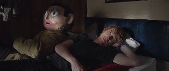 Movie still from “Frank” (2014), directed by Lenny Abrahamson – A man laying in a bed next to a creepy doll; Medium shot, Over the shoulder angle