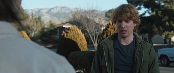 Movie still from “Frank” (2014), directed by Lenny Abrahamson – A young man standing in front of some bushes; Medium shot, Over the shoulder angle