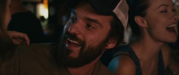 Movie still from “Drinking Buddies” (2013), directed by Joe Swanberg – A man with a beard wearing a hat and smiling; Close Up shot, Over the shoulder angle