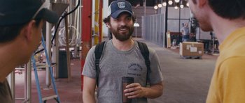 Movie still from “Drinking Buddies” (2013), directed by Joe Swanberg – A man with a beard wearing a hat and holding a cup; Medium shot, Over the shoulder angle