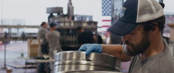 Movie still from “Drinking Buddies” (2013), directed by Joe Swanberg – A person in a blue hat and blue gloves is holding a keg; Close Up shot, Over the shoulder angle