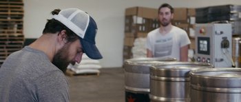 Movie still from “Drinking Buddies” (2013), directed by Joe Swanberg – A man in a baseball cap and a man in a white t - shirt are in a warehouse; Close Up shot, Over the shoulder angle