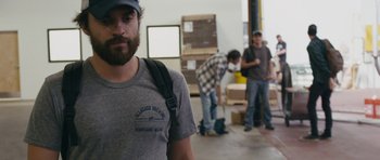 Movie still from “Drinking Buddies” (2013), directed by Joe Swanberg – A man with a beard wearing a hat and a gray shirt; Medium shot, Over the shoulder angle