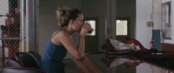 Movie still from “Drinking Buddies” (2013), directed by Joe Swanberg – A woman sitting at a table drinking from a cup; Medium shot, Over the shoulder angle