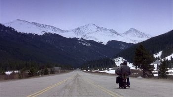 Movie still from “Dumb and Dumber” (1994), directed by Peter Farrelly – A person riding a motorcycle down a road with mountains in the background; Extreme Wide shot, High angle
