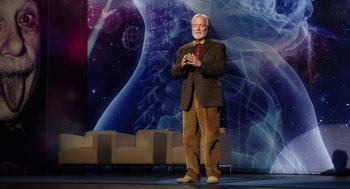 Movie still from “Dumb and Dumber To” (2014), directed by Peter Farrelly – An older man standing on a stage in front of an image of the earth; Medium shot, Low angle