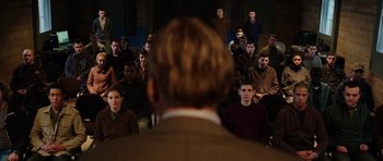 Movie still from “Dying of the Light” (2014), directed by Paul Schrader – A group of people sitting in a room with a man in front of them; Medium shot, Over the shoulder angle