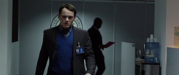 Movie still from “Dying of the Light” (2014), directed by Paul Schrader – A man in a suit and tie standing in front of a wall; Medium shot, Over the shoulder angle