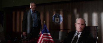 Movie still from “Dying of the Light” (2014), directed by Paul Schrader – A man standing in front of an american flag in a room; Medium shot, Over the shoulder angle