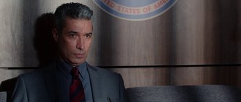 Movie still from “Dying of the Light” (2014), directed by Paul Schrader – A man wearing a suit and tie in front of the seal of the united states of america; Close Up shot, Over the shoulder angle