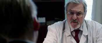 Movie still from “Dying of the Light” (2014), directed by Paul Schrader – A man wearing a white lab coat and glasses talking to another man; Close Up shot, Over the shoulder angle