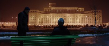 Movie still from “Dying of the Light” (2014), directed by Paul Schrader – A person sitting on a bench in front of a large building; Extreme Wide shot, Low angle