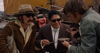 Movie still from “Easy Rider” (1969), directed by Dennis Hopper – A group of men standing next to each other eating food; Medium shot, Over the shoulder angle