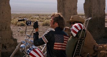 Movie still from “Easy Rider” (1969), directed by Dennis Hopper – A man riding on the back of a motorcycle with american flags on it; Medium shot, Low angle