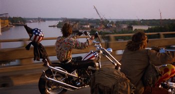 Movie still from “Easy Rider” (1969), directed by Dennis Hopper – A man riding on the back of a motorcycle on a bridge; Wide shot, Over the shoulder angle