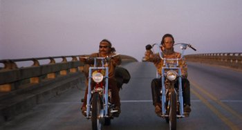 Movie still from “Easy Rider” (1969), directed by Dennis Hopper – Two people riding motorcycles on a road with a bridge in the background; Wide shot, Low angle