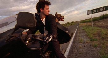 Movie still from “El Mariachi” (1992), directed by Robert Rodriguez – A man riding a motorcycle with a dog on the back of it; Wide shot, Low angle