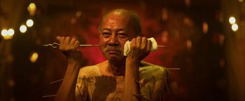 Movie still from “Elephant White” (2011), directed by Prachya Pinkaew – An older man holding a stick with food on it; Close Up shot, Low angle