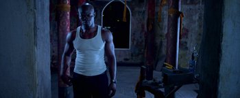 Movie still from “Elephant White” (2011), directed by Prachya Pinkaew – A man in a white tank top standing next to a window; Medium shot, Low angle