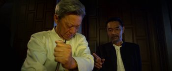 Movie still from “Elephant White” (2011), directed by Prachya Pinkaew – A man in a priest's outfit is holding a fork; Medium shot, Over the shoulder angle