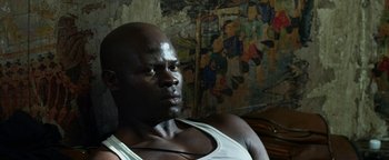 Movie still from “Elephant White” (2011), directed by Prachya Pinkaew – A black man with a white tank top is sitting in front of a mural; Close Up shot, High angle