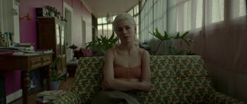 Movie still from “Ema” (2019), directed by Pablo Larraín – A woman sitting on top of a couch with her arms crossed; Medium shot, Low angle