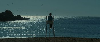 Movie still from “Ema” (2019), directed by Pablo Larraín – A woman sitting on a lifeguard chair looking out at the ocean; Extreme Wide shot, High angle