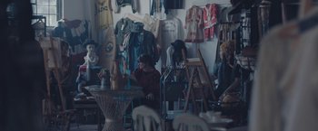 Movie still from “Endings, Beginnings” (2019), directed by Drake Doremus – Two people sitting at a table in a room with a lot of clothes hanging on the wall; Wide shot, High angle