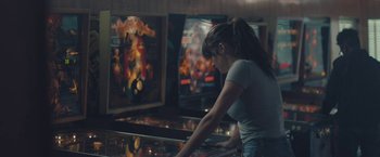 Movie still from “Endings, Beginnings” (2019), directed by Drake Doremus – A woman standing in front of a pinball machine; Medium shot, Over the shoulder angle