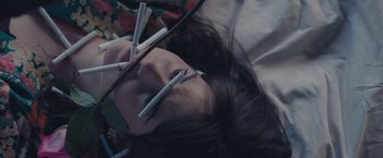 Movie still from “Endings, Beginnings” (2019), directed by Drake Doremus – A woman laying on the ground holding a pair of cigarette lighters in her mouth; Extreme Close Up shot, Overhead angle