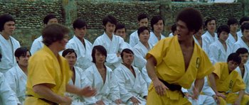 Movie still from “Enter the Dragon” (1973), directed by Robert Clouse – A group of men in white robes sitting next to each other in front of a stone wall; Medium shot, Over the shoulder angle