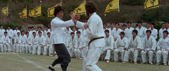 Movie still from “Enter the Dragon” (1973), directed by Robert Clouse – Two men in white shirts are practicing martial arts in front of a group of people; Medium shot, Over the shoulder angle