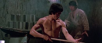 Movie still from “Enter the Dragon” (1973), directed by Robert Clouse – A shirtless man holding a wooden stick in a room; Medium shot, Low angle