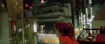 Movie still from “Enter the Void” (2009), directed by Gaspar Noé – A woman with red hair is standing in front of a building at night; Medium shot, Over the shoulder angle