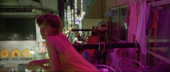 Movie still from “Enter the Void” (2009), directed by Gaspar Noé – A woman in a pink dress sitting on a chair in front of a window; Medium shot, Over the shoulder angle