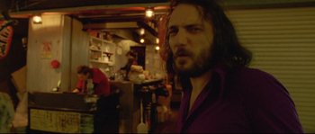 Movie still from “Enter the Void” (2009), directed by Gaspar Noé – A man with long hair and a purple shirt is in a room; Medium shot, Low angle