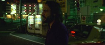 Movie still from “Enter the Void” (2009), directed by Gaspar Noé – A man with long hair and beard standing on the side of the street; Medium shot, Over the shoulder angle