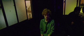 Movie still from “Enter the Void” (2009), directed by Gaspar Noé – A man with curly hair standing in a dark room; Medium shot, Over the shoulder angle