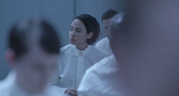 Movie still from “Equals” (2015), directed by Drake Doremus – A woman in a white shirt is sitting in front of a group of people; Medium shot, Over the shoulder angle