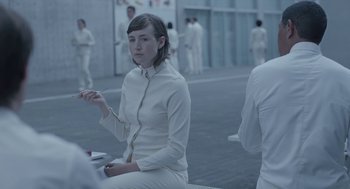 Movie still from “Equals” (2015), directed by Drake Doremus – A woman in all white sitting on a bench in front of a group of people; Medium shot, Over the shoulder angle