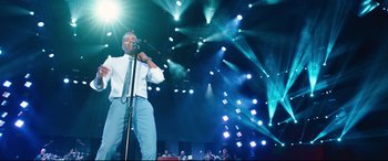 Movie still from “Girls Trip” (2017), directed by Malcolm D. Lee – A man in a white dress shirt and tie holding a microphone on a stage; Medium shot, Low angle