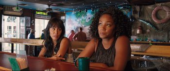 Movie still from “Girls Trip” (2017), directed by Malcolm D. Lee – Two women sitting at a table in front of a painting; Medium shot, Over the shoulder angle