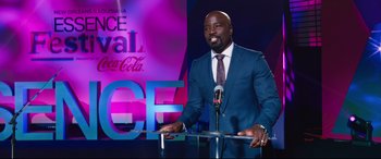 Movie still from “Girls Trip” (2017), directed by Malcolm D. Lee – A man standing at a podium in front of a microphone; Medium shot, Over the shoulder angle