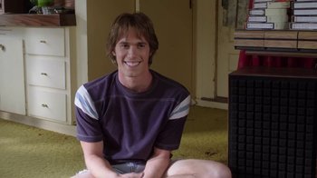 Movie still from “Everybody Wants Some!!” (2016), directed by Richard Linklater – A man sitting on the ground smiling for the camera; Medium shot, Low angle