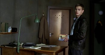 Movie still from “Evil” (2003), directed by Mikael Håfström – A young man holding a book standing in front of a desk; Medium shot, Over the shoulder angle
