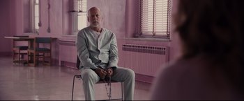 Movie still from “Glass” (2019), directed by M. Night Shyamalan – An older man sitting on a chair with chains around his ankles; Medium shot, Over the shoulder angle