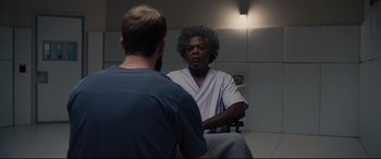 Movie still from “Glass” (2019), directed by M. Night Shyamalan – A man in a hospital gown is talking to another man in a hospital room; Medium shot, Over the shoulder angle