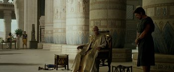 Movie still from “Exodus: Gods and Kings” (2014), directed by Ridley Scott – A man sitting on a chair in a room with pillars; Wide shot, Low angle