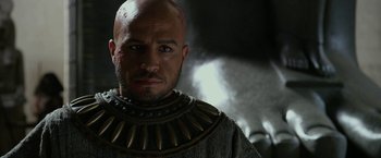 Movie still from “Exodus: Gods and Kings” (2014), directed by Ridley Scott – A bald man wearing a gold necklace and a black shirt; Close Up shot, Over the shoulder angle