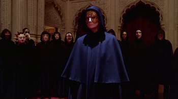 Movie still from “Eyes Wide Shut” (1999), directed by Stanley Kubrick – A man in a blue robe and mask stands in front of a group of people; Wide shot, Low angle